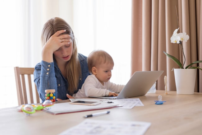 trött arbetande mamma framför dator och dokument med barn i knät